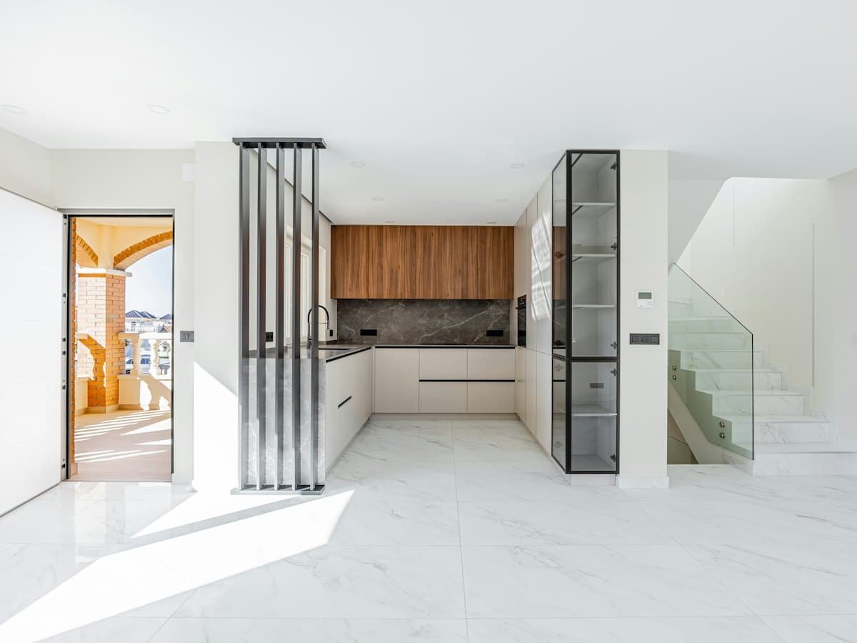 A modern kitchen with wooden cabinets and marble floors opening onto a bright living area.